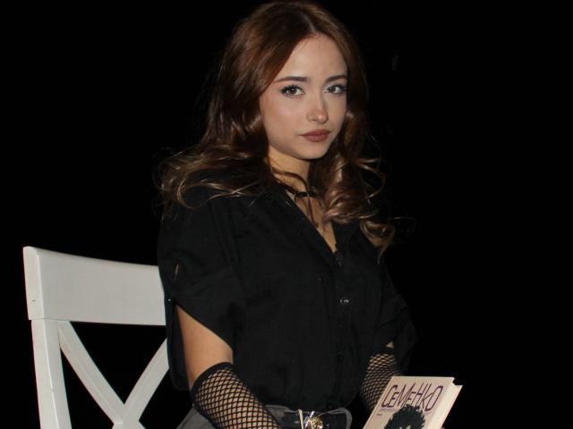 EmiliaMuse con cabello ondulado castaño, camisa negra, guantes de rejilla, silla blanca, sosteniendo un papel, fondo negro, expresión seria, plano medio, senos visibles, complexión delgada, asiática..