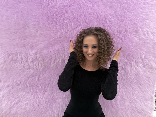 MichelleHotX69 with curly brown hair, smiling, wearing a black long-sleeve top, hands raised, standing in front of a pink fluffy wall. Boobs are not visible.