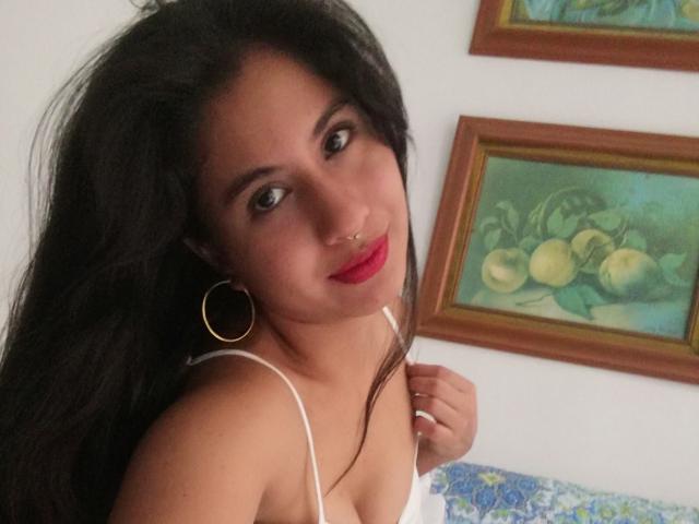 AuroraMonroe has dark hair, red lipstick, and hoop earrings, with a nose ring, looking at the camera, wearing a white strap dress, showing her tits.