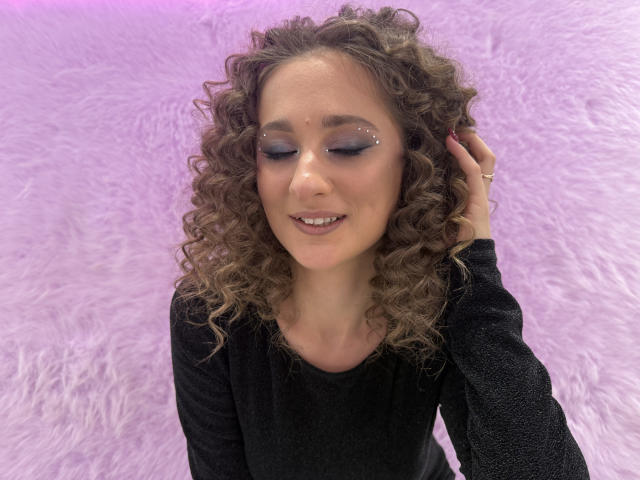 MichelleHotX69 has curly brown hair, black sparkly eye makeup, and a cute smile, wearing a black long-sleeve shirt, with one hand in her hair.