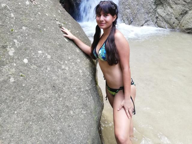 AuroraMonroe with long brown pigtails, wearing a green and blue patterned bikini, standing by a rock, water flowing in the background, smiling, hands on rock, legs in water.