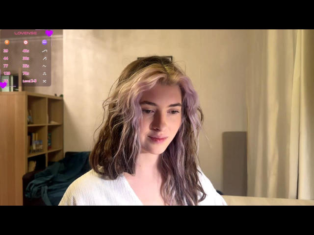 MaryamRisby with wavy, purple and brown hair, white shirt, and subtle smile, looking down with soft lighting, in a slightly messy room. Her tits are small and cute.