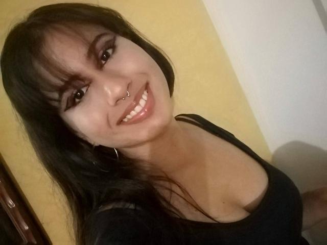 AuroraMonroe with long black hair, brown eyes, nose ring, and hoop earrings, smiling at the camera in a black top, showing her boobs in a selfie.