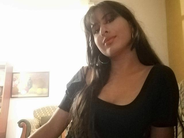 AuroraMonroe with long black hair, hoop earrings, and a black tee, slightly pouting with a subtle smile, showing her tits, in a warmly lit room.