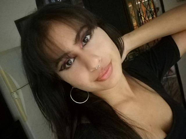AuroraMonroe has black hair, blue eyes, and a nose ring. She's wearing a black top, hoop earrings, and is smiling slightly with her arm raised. Her tits are small.