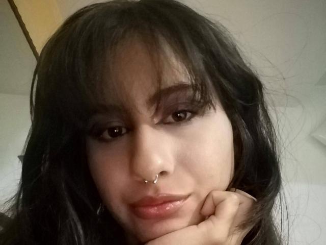 AuroraMonroe with dark, wavy hair, brown eyes, and a nose ring, resting her chin on her hand, looking at the camera with a slight, sexy smile.