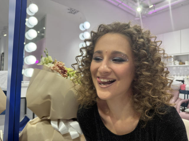 MichelleHotX69 with curly brown hair, black glittery top, blue eyeshadow, smiling, holding brown paper bouquet, white ribbon, in brightly lit room with round mirrors.