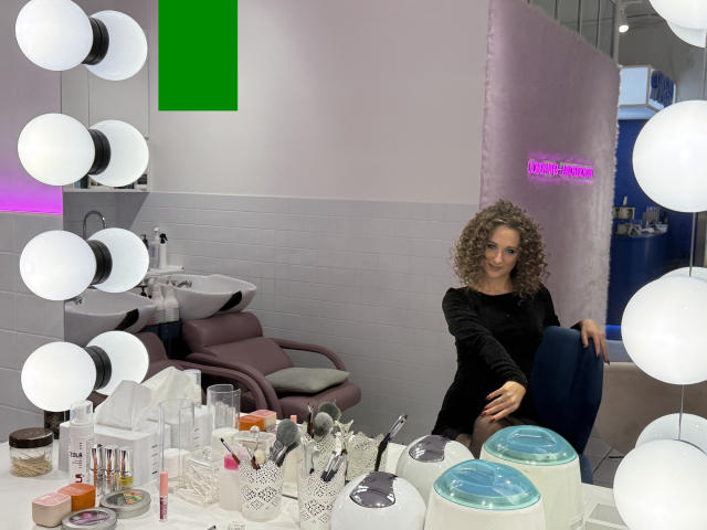 MichelleHotX69 with curly brown hair, wearing a black long-sleeve top, sitting in a makeup room with round white lights, makeup tools, and containers on a white table.