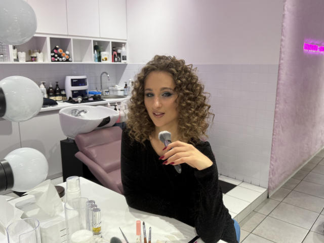 MichelleHotX69 with curly brown hair, wearing a black long-sleeve top, holding a makeup brush, sitting at a white countertop with various makeup items and a pink chair.