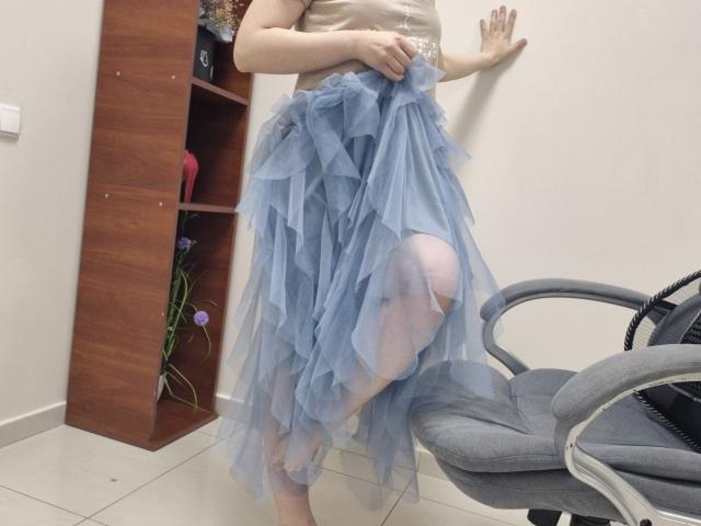 JoannaSia has a blue, ruffled, layered skirt, exposing her pale, bare thigh and knee. She's wearing a shiny, beige top, standing in front of a wooden shelf.