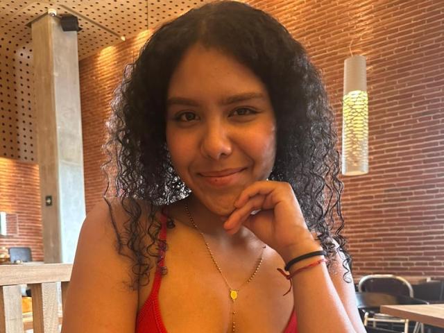 AriaVega with curly black hair, red bra, smiling, hand on chin, gold necklace, brown brick wall, warm lighting, and wooden ceiling, in a casual cafe setting.
