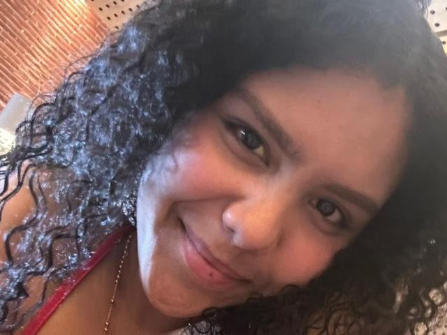 AriaVega has curly black hair, brown eyes, and a slight smile with a small red blemish on her chin. She's wearing a red top with a gold necklace.