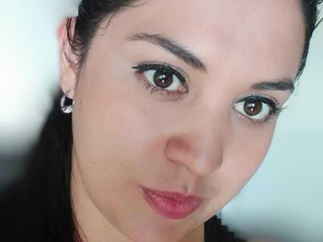 PaulaAnd's face with brown eyes, black eyeliner, dark eyebrows, pink lipstick, slightly pouted lips, and black hair pulled back. She's wearing small hoop earrings.