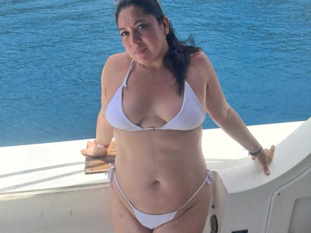 PaulaAnd is a curvy woman with white bikini, small boobs, and a slightly chubby ass, standing on a boat, smiling, with blue water in the background.