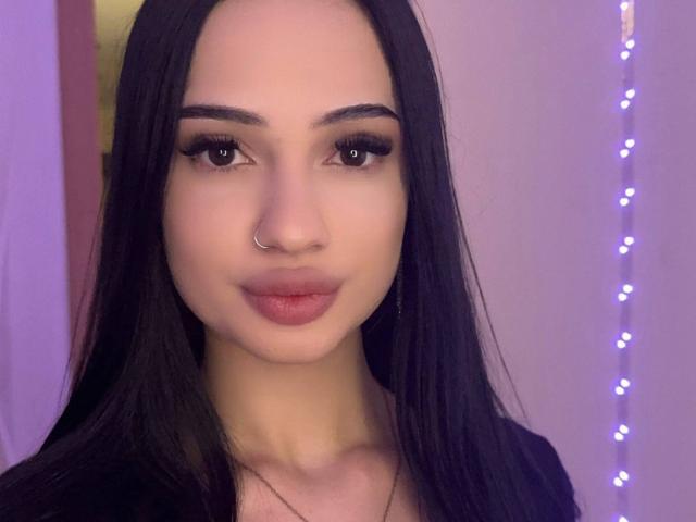 ShirleySiegel with long black hair, nose ring, and full lips, looking at the camera with dark, thick eyebrows and long, thick eyelashes. Soft purple lighting in the background.