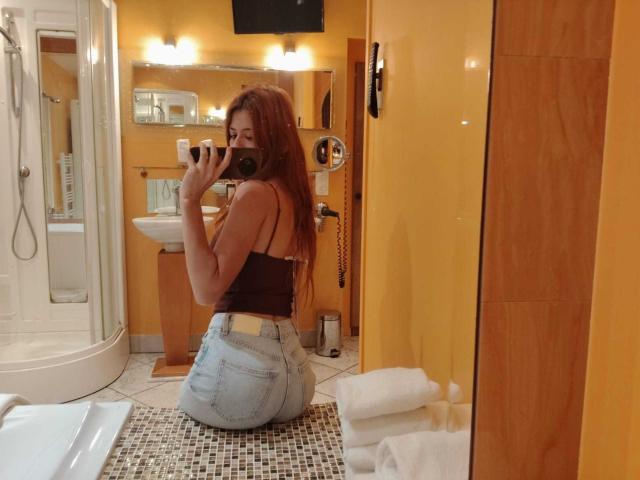JuliethLove with long red hair, wearing a black tank top and high-waisted blue jeans, kneeling, taking a mirror selfie, showing her ass, in a yellow-tiled bathroom.