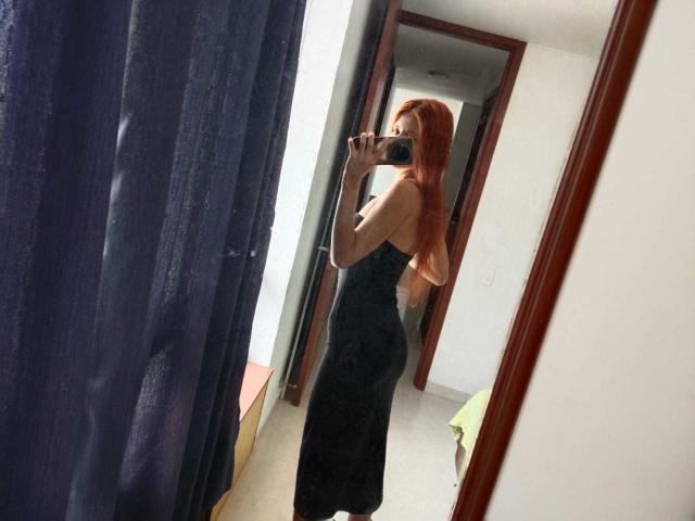 JuliethLove with red hair, wearing a black strapless dress, showing her ass in a mirror selfie, holding a phone, in a room with dark curtains.