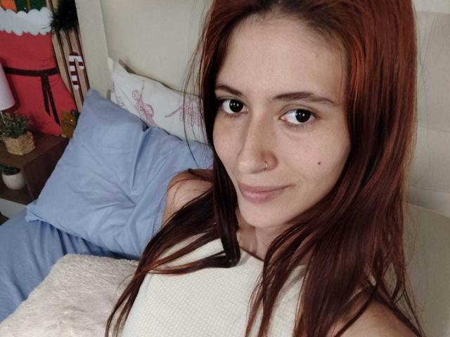 JuliethLove with red hair, small nose ring, and light skin, smiling at the camera, wearing a white sleeveless top, lying in bed with blue pillows.