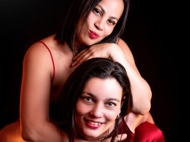 SaharaMK with dark brown hair, red lipstick, and red dress, arms around another SaharaMK with similar hair and red lipstick, smiling, both have visible tits, black background.