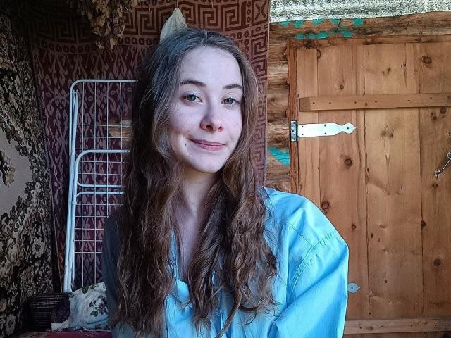 EmileeBaggerly with wavy brown hair, light skin, wearing a blue shirt, smiling subtly, standing in front of a wooden door with a white latch.
