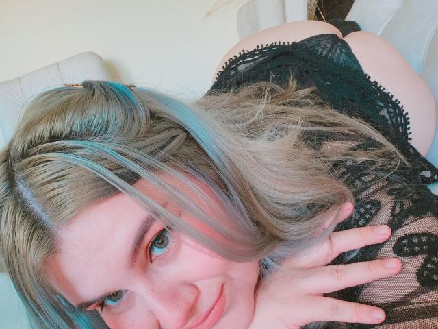 Akkanee with blonde and blue highlights, smiling, wearing black lace lingerie, bending over, showing her ass, hands near face, on a white couch.