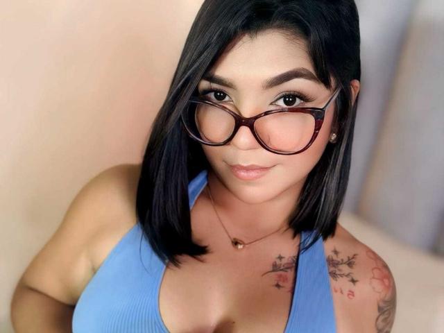 ValeryPierce with black bob, glasses, blue tank top, tattoos on shoulders, smiling, medium tits, light skin, brown eyes, subtle makeup, small nose, pink lips, chest necklace.
