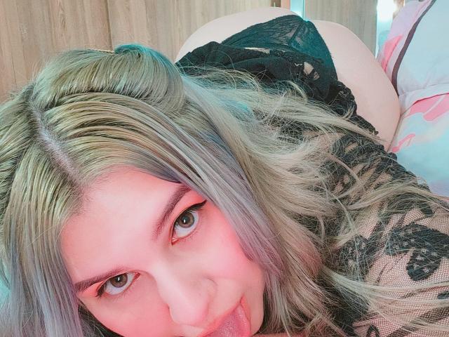 Akkanee with blonde hair, green eyes, and pink eyeliner, tongue out, lying on a bed, wearing black lace lingerie, showing her ass. Her makeup and hair are slightly messy.