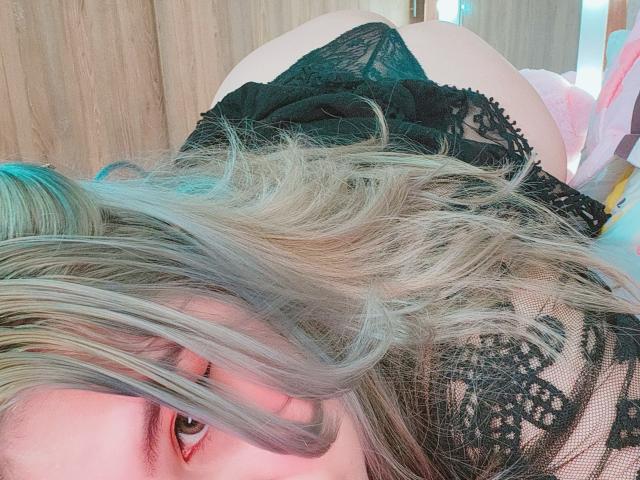 Akkanee's face is close to the camera, lying on her back with blonde and blue-tipped hair, wearing black lace panties, showing her ass. Her eyes are looking up.