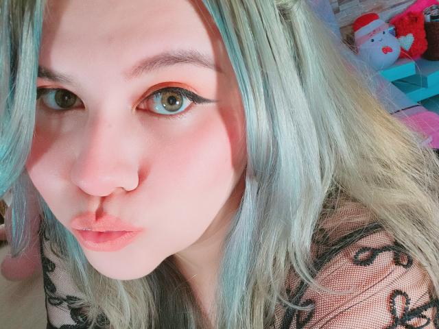 Akkanee has pale skin, green eyes, blue-green hair, and pink lips. She's wearing black lace and has a nose ring. Her makeup includes orange eyeshadow.