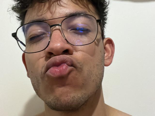 MiloJhones, con labios carnosos, lleva gafas, tiene el cabello oscuro ligeramente despeinado y una barba clara, con una expresión neutra y un sutil reflejo de luz azul en sus ojos..