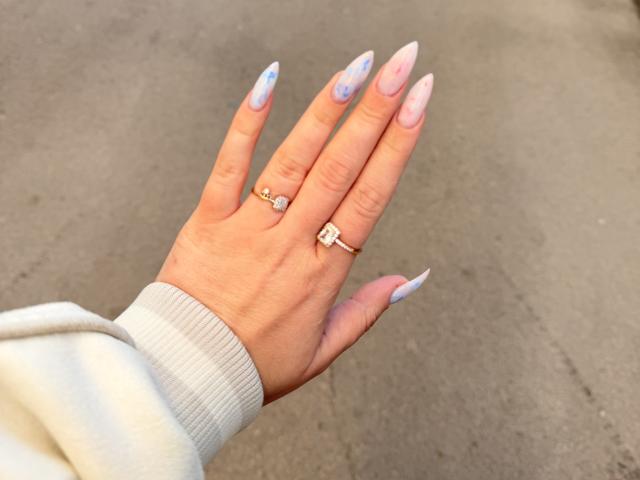 SugarrX's hand with pink and blue ombre nails, wearing a gold ring with a diamond and a smaller ring with a heart symbol, in a white sleeve.