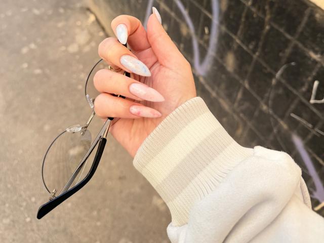 SugarrX's hand with long, pink, almond-shaped nails holding black glasses with clear lenses. She's wearing a white, striped, long-sleeve sweater.