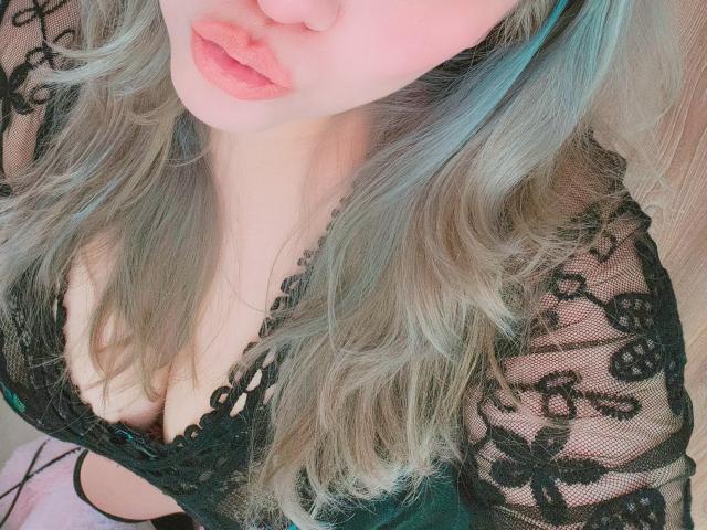 Akkanee's face and upper body, showing pink lips and a black lace top with floral patterns, highlighting her cleavage and wavy, blue-green hair.