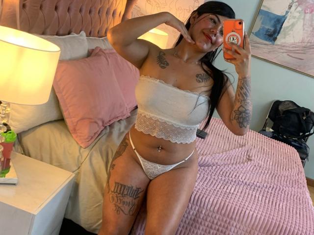 CatalinaReiyna in white lace lingerie, showing off her tits and pussy, with tattoos on her chest and thigh, taking a mirror selfie in a pink-bedroom.