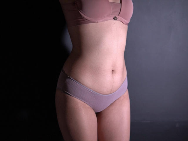 AliCipher's pink bra and purple panties show her curvy, pale-skinned body with small tits and a slight belly, standing in dim light. Her ass is partially visible.