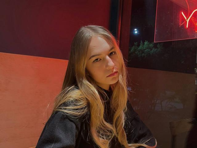 NenitaTraino con cabello rubio largo, vistiendo una camisa negra, mirando a la cámara con una expresión seria, iluminación roja en el fondo, y una señal de neón parcialmente visible..