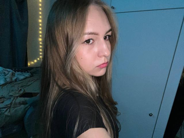NenitaTraino mit langen blonden Haaren, heller Haut und rosafarbenen Lippen, trägt ein schwarzes Hemd und blickt mit ernstem Gesichtsausdruck über ihre Schulter, in einem schwach beleuchteten Raum..