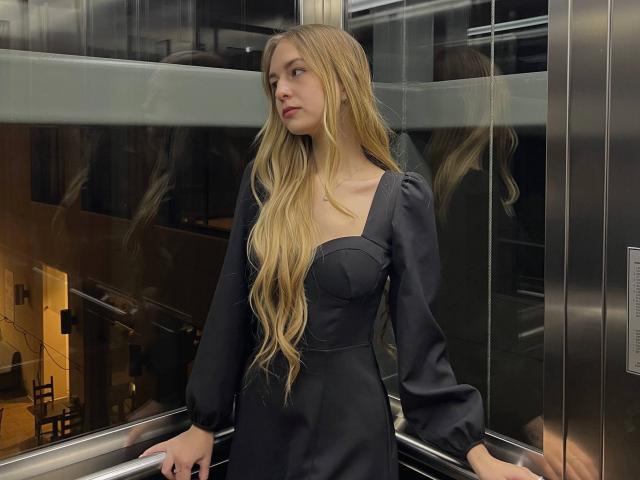 NenitaTraino, avec de longs cheveux blonds, portant une robe noire à manches bouffantes, se tient dans un ascenseur, regardant sur le côté, les mains sur la rambarde, reflet dans le verre..