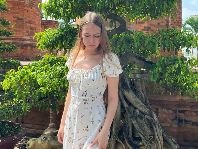 NenitaTraino, blonde, portant une robe blanche à fleurs, se tient devant un grand arbre aux racines noueuses, le regard baissé, petite poitrine, mignonne, mince, peau claire, journée ensoleillée, en plein air..