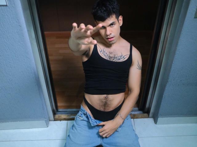 DerekVoss, com cabelo preto curto, vestindo uma regata preta, expondo um peito tatuado, roupa íntima preta e jeans abotoados, aponta para frente com a mão direita..