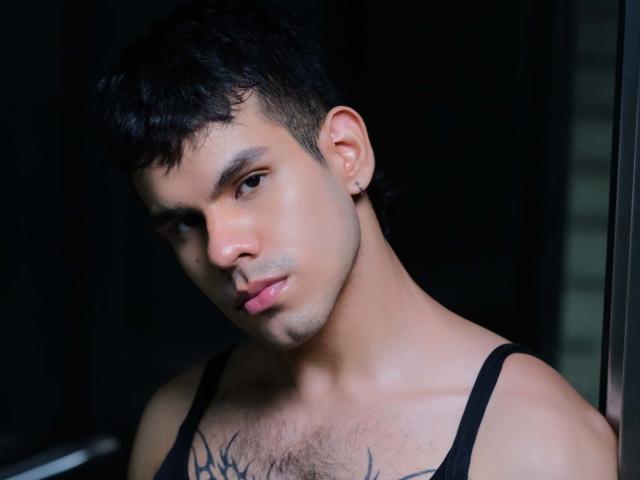 DerekVoss with short black hair, dark eyebrows, ear piercing, slight chest hair, black tank top, and tattoo on chest, looking down with a serious expression.