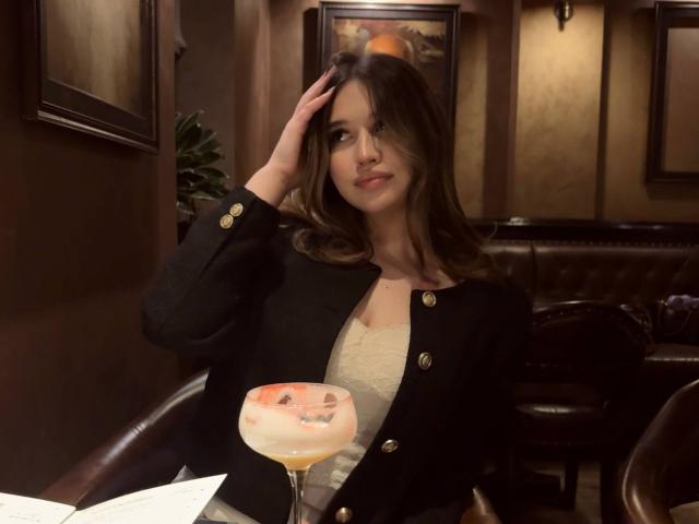 LaylaMantsch with long brown hair, wearing a black jacket with gold buttons, holding a pink cocktail, sitting in a dimly lit bar, hand in hair, slight smile.
