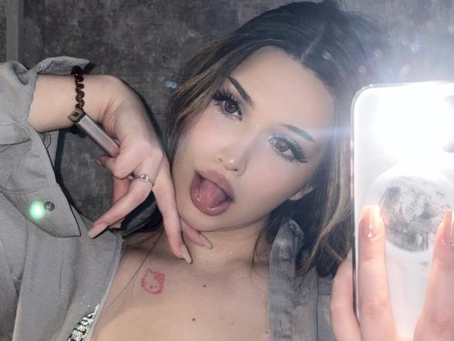 LaylaMantsch with tongue out, makeup on, holding phone, gray jacket, brown bracelet, ring, small tattoo near collarbone, mirror selfie, reflection visible. Boobs slightly visible, long brown hair.