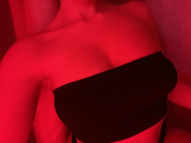 Red-lit photo of OliviaGranda's bare shoulders and black strapless top, highlighting her large, round boobs under soft, warm lighting. Her skin is smooth and slightly shiny.
