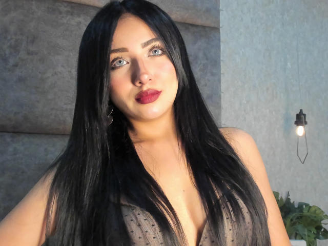 LillyTaylor with long black hair, blue eyes, and red lipstick, wearing a sheer black top, showing her tits, standing in front of a gray wall with a light bulb.