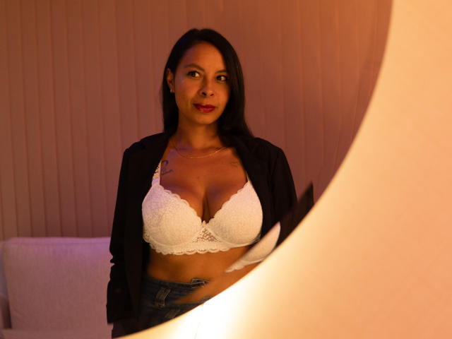 ClairBennet with medium brown skin, black hair, wearing a white lace bra, black cardigan, and blue jeans, standing in front of a warm, orange-lit, circular mirror.