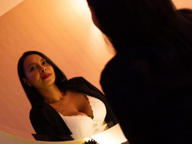 Clair Bennet, met donker haar, rode lippenstift en een zwarte blazer, die haar grote witte borsten laat zien, kijkt omhoog naar iemand met lang donker haar op de voorgrond..