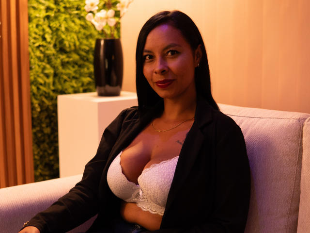 ClairBennet with dark hair, red lipstick, wearing a black blazer over a white lace bra, showing her big tits, sitting on a white chair, smiling softly.