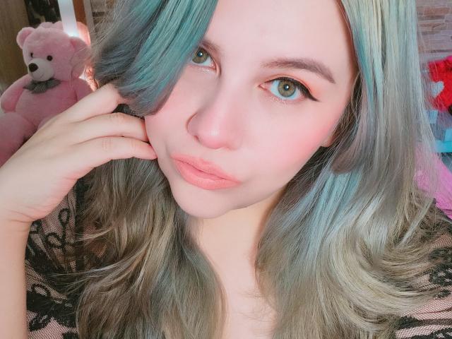Akkanee has pale skin, blue-green hair, and pink lipstick, with her hand near her face, green eyes, and wearing a black lace top, looking at the camera.
