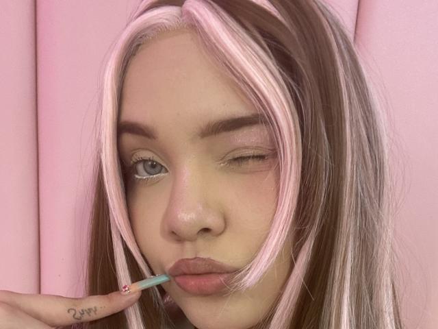 AlayaWillow with pink and brown hair, winking, applying pink lipstick with a blue pencil, has a tattoo on her right ring finger, pink background.